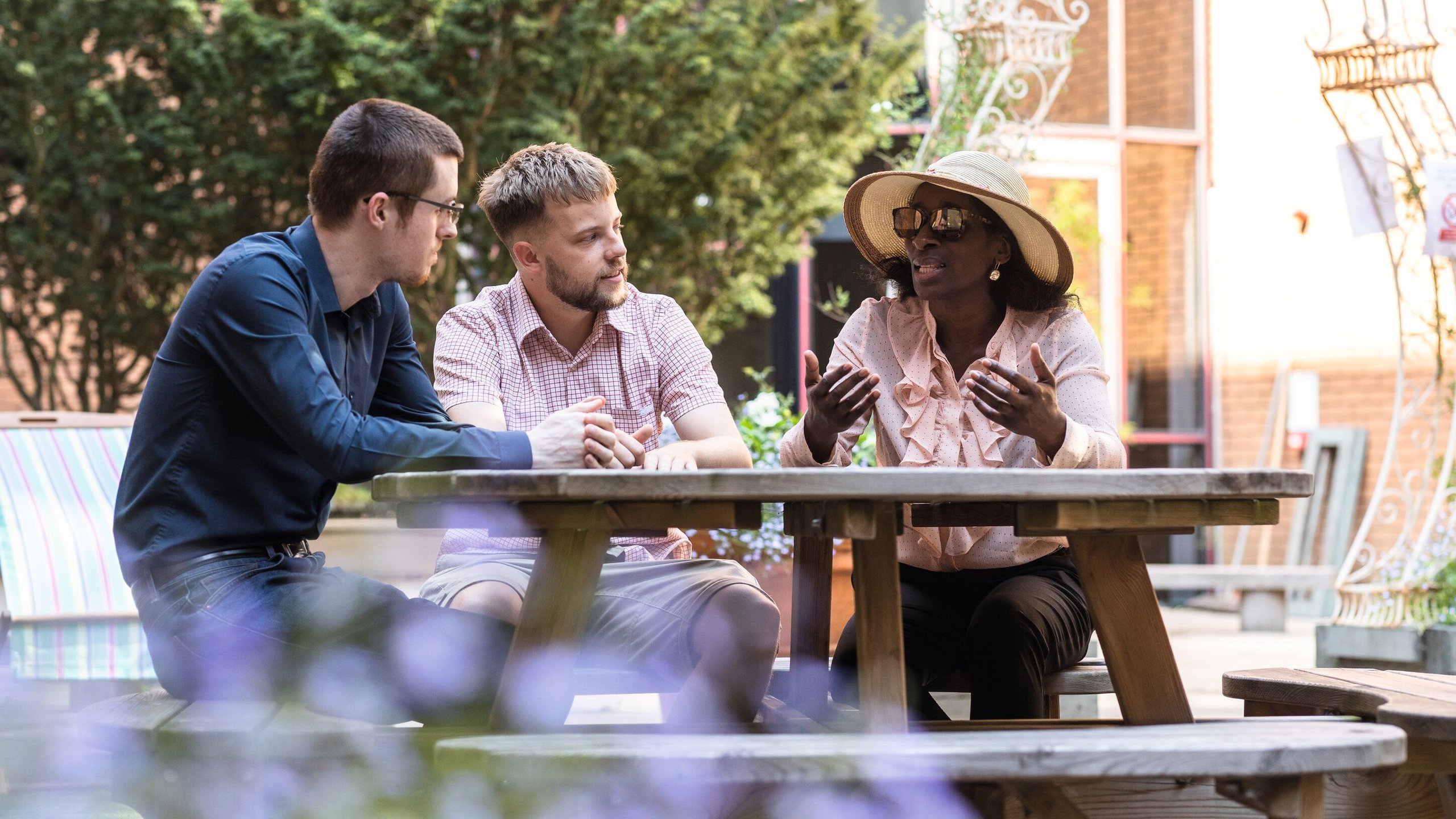 Three colleagues, two men and a woman in a hat and sunglasses, sit at a picnic table outdoors, engaged in conversation.