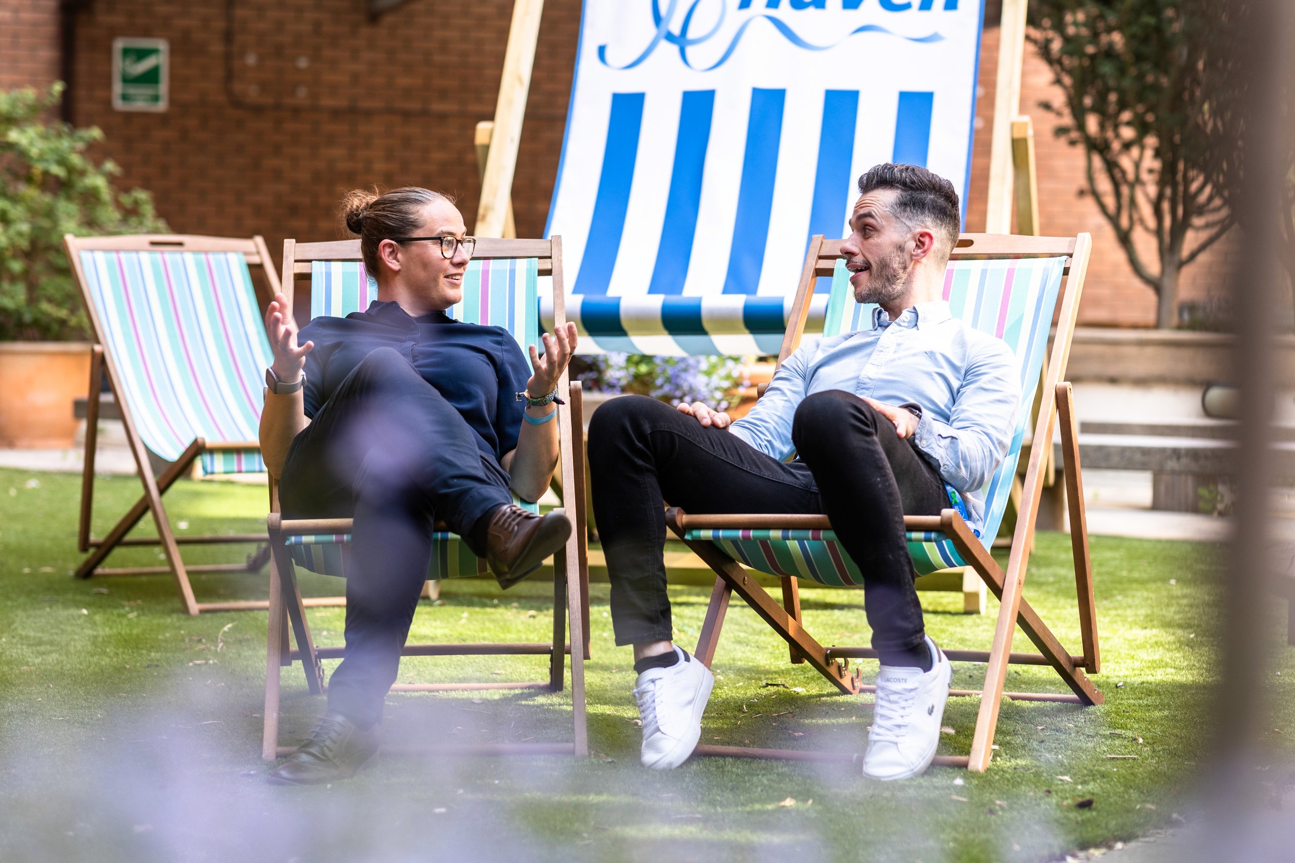 Two men in business casual attire sit in deck chairs on a lawn, talking.