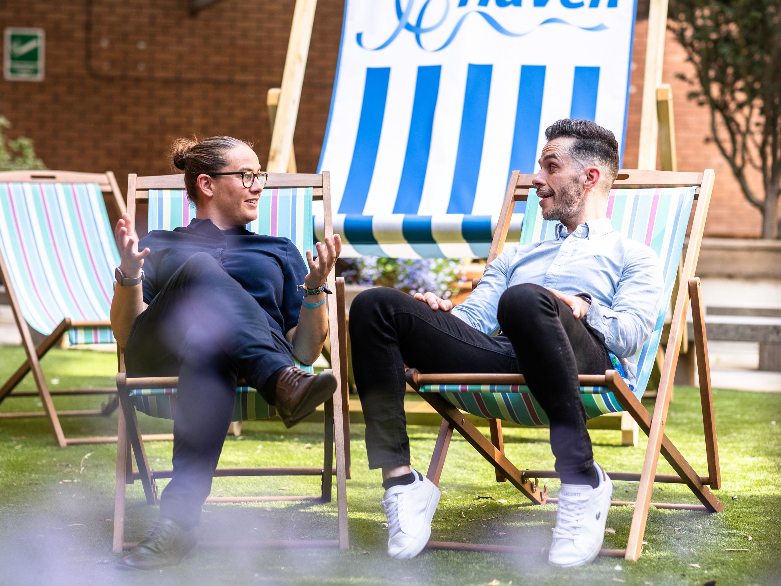 Two men in business casual attire sit in deck chairs on a lawn, talking.
