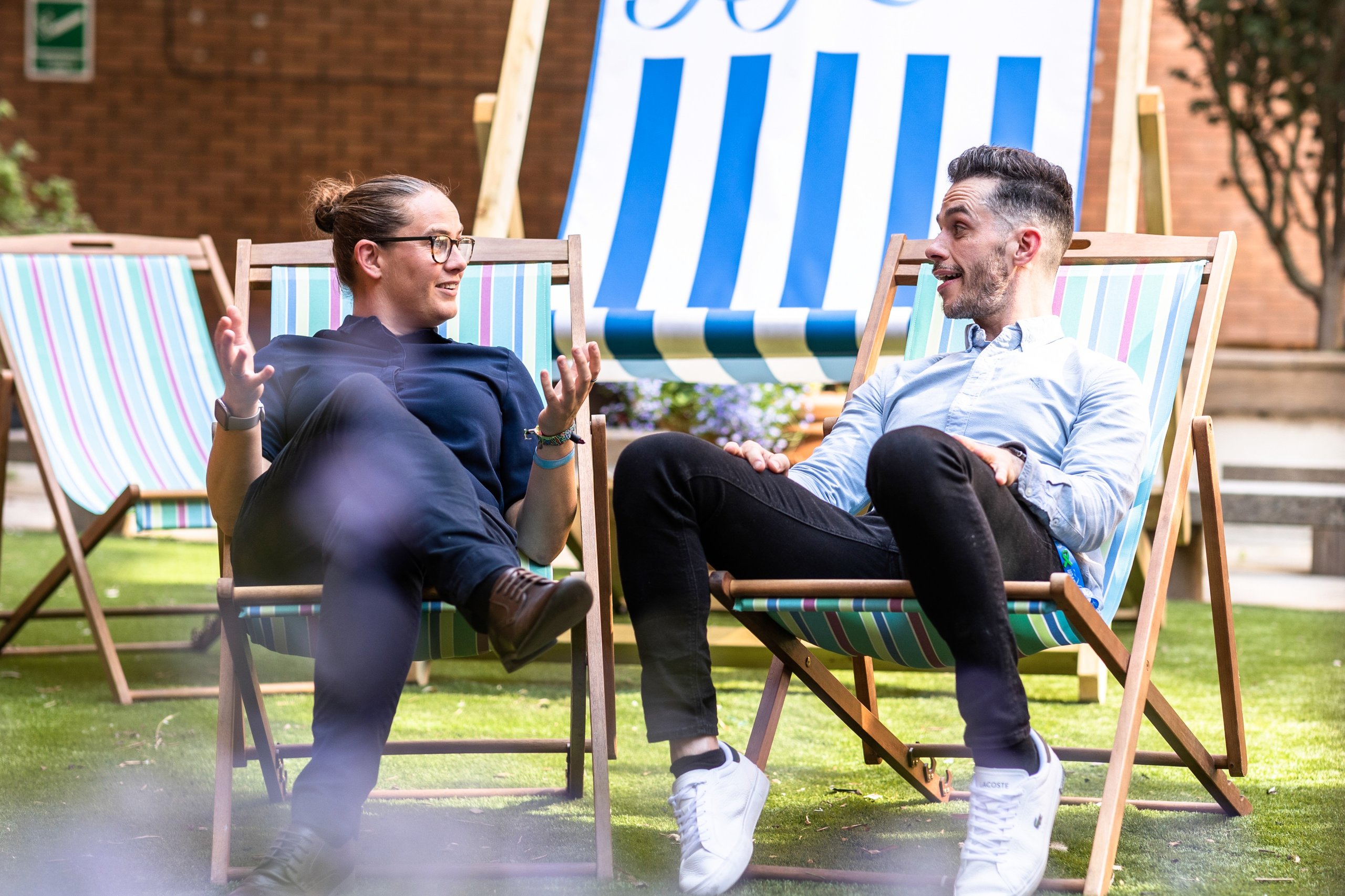 Two men in business casual attire sit in deck chairs on a lawn, talking.