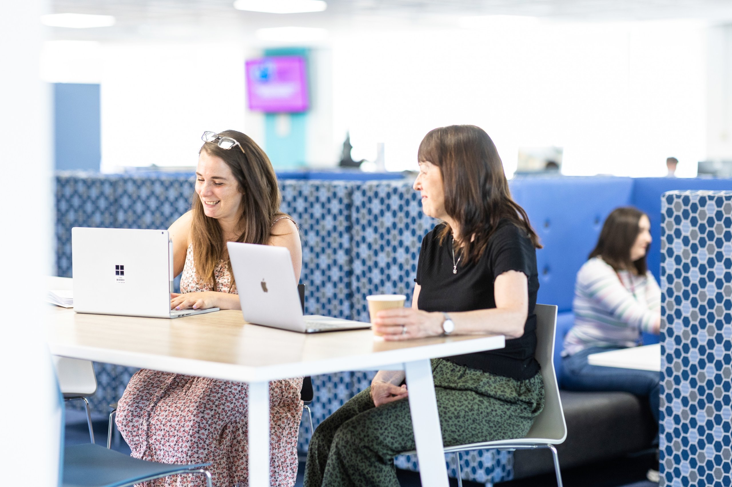 Two women work on laptops at a table in a modern office space, with another person visible in the background.