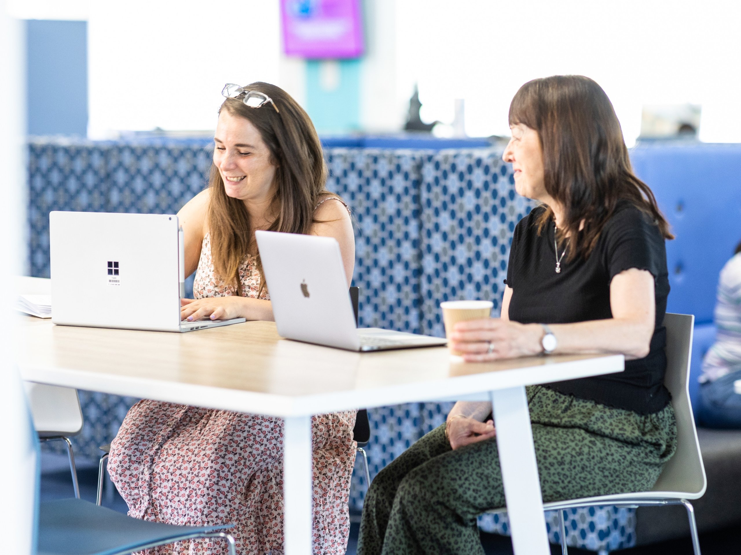 Two women work on laptops at a table in a modern office space, with another person visible in the background.