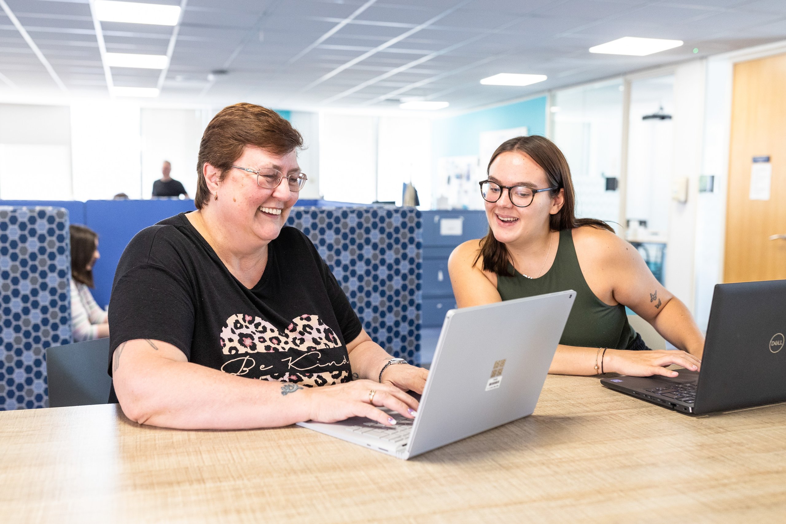Two women of different ages are smiling and working together on laptops at a wooden desk in a modern office.