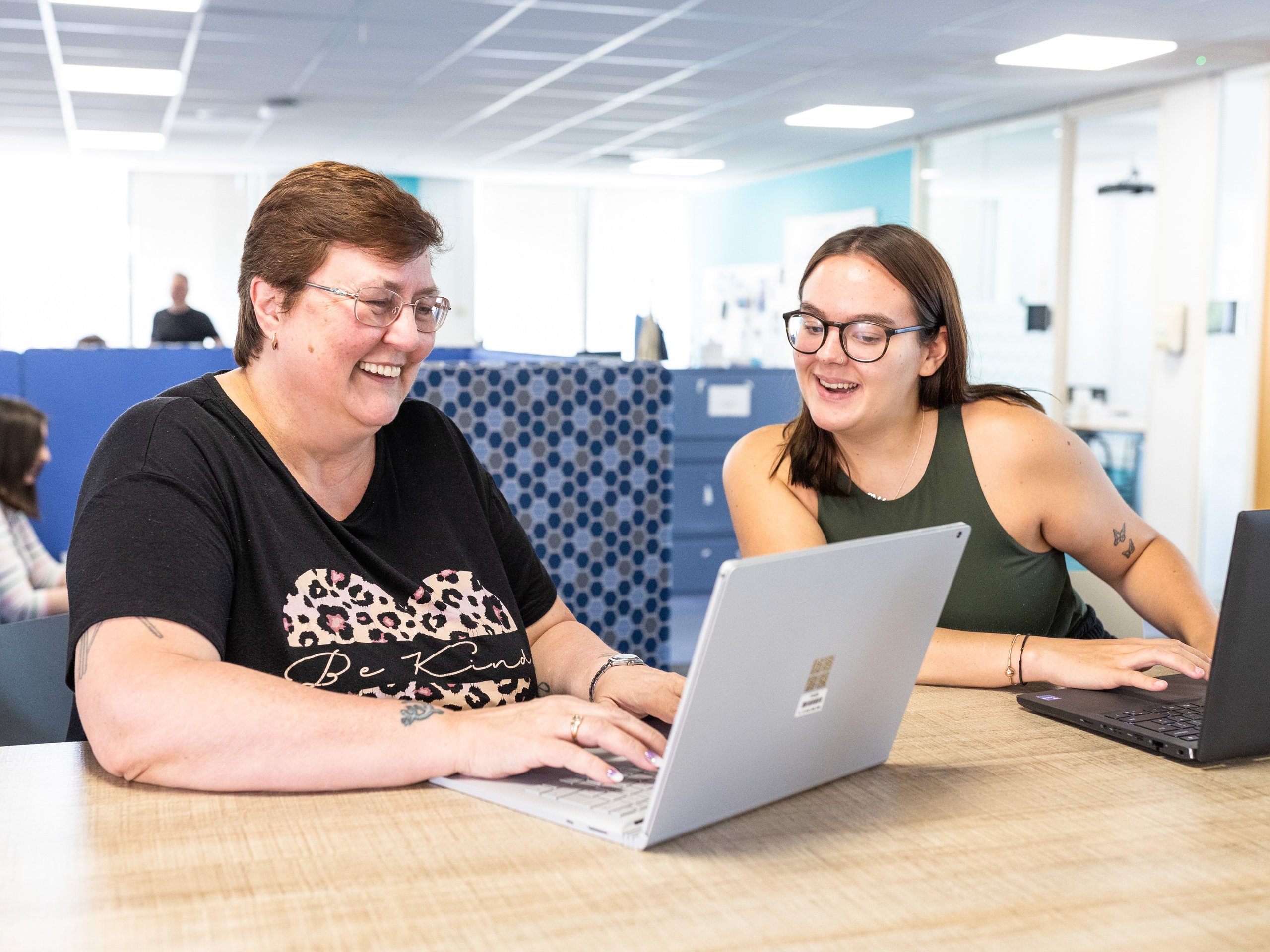 Two women of different ages are smiling and working together on laptops at a wooden desk in a modern office.