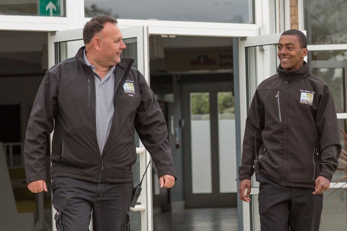 Two security guards in matching black jackets and pants walk out of double glass doors. One guard wears a grey shirt, the other a black shirt.