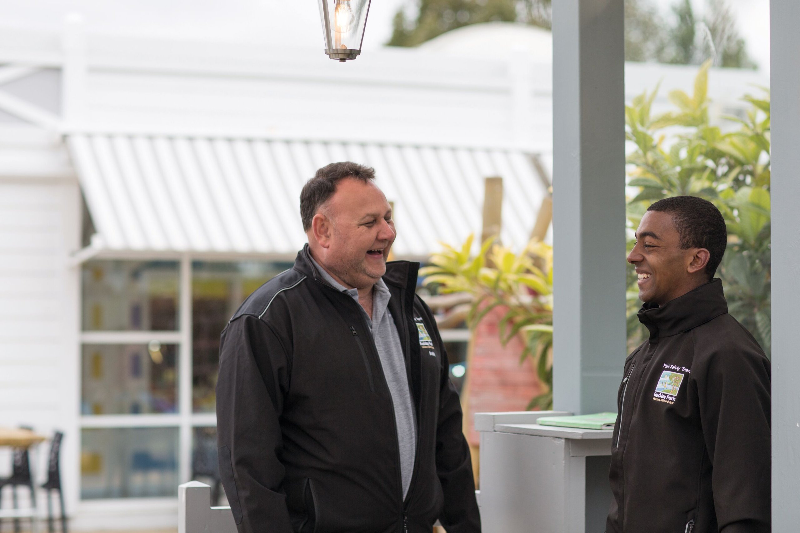 Two men in park safety uniforms laugh together outside a building with a striped awning.