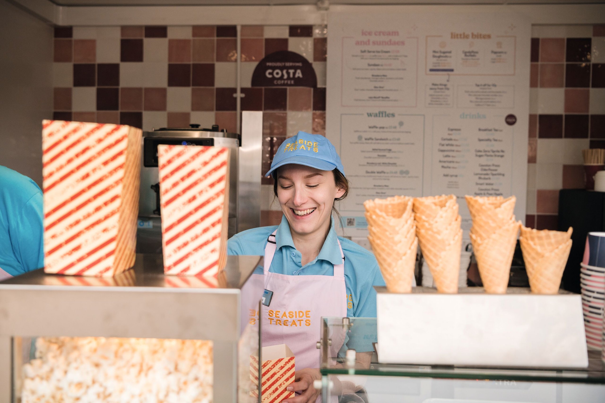 Employee wearing a blue shirt and pink apron smiles while standing behind a counter with popcorn, ice cream cones, and a menu.