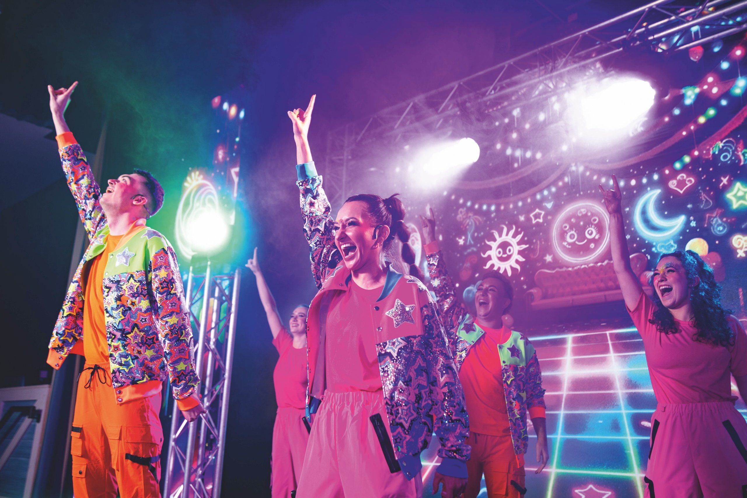 Dancers in brightly colored, star-patterned jackets and orange or pink outfits perform on a brightly lit stage with a neon backdrop.