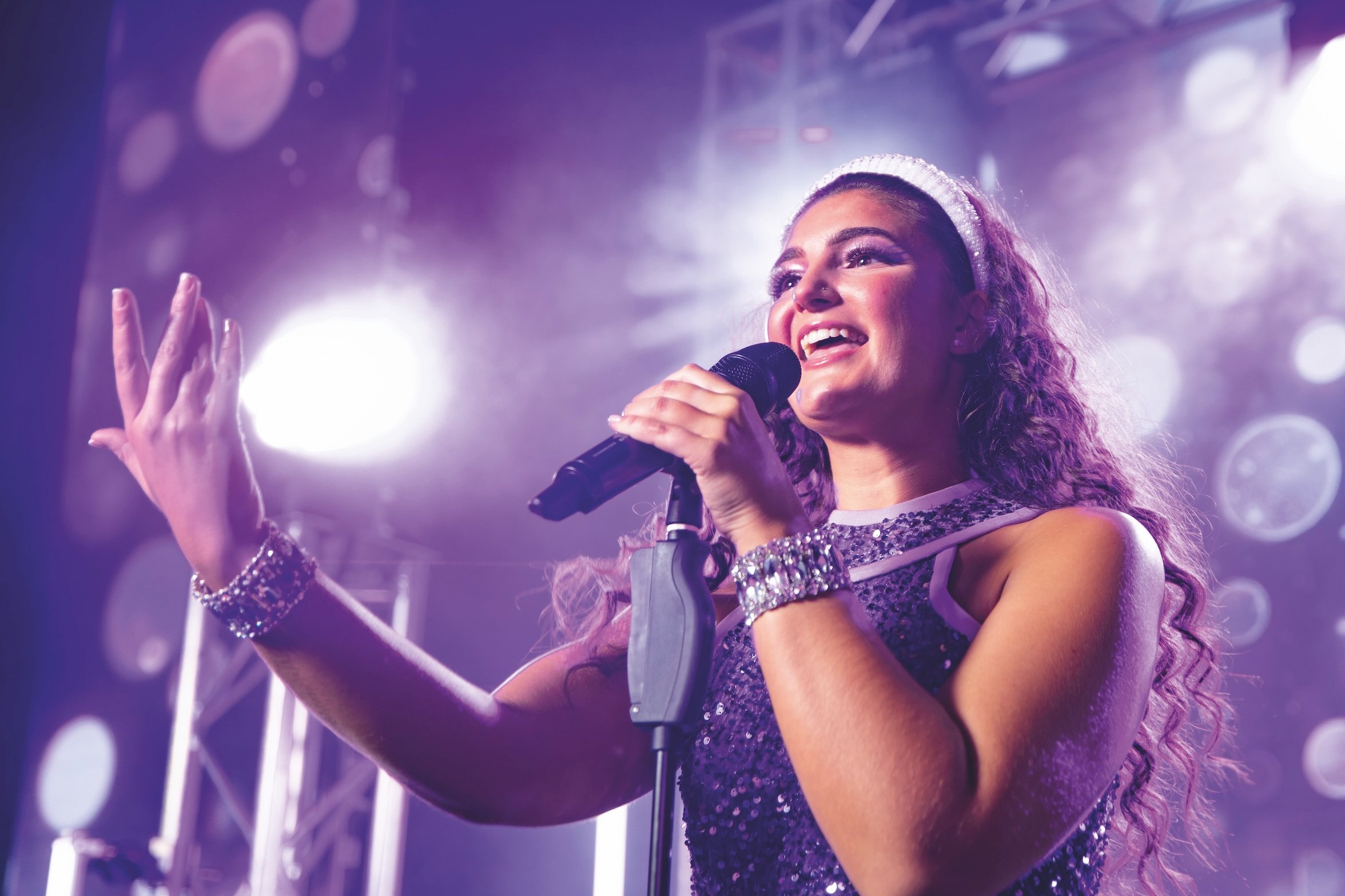 A female singer in a sequined outfit and headband sings into a microphone on a stage with purple lighting and bokeh effects.