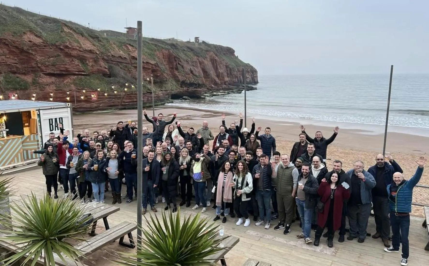 A large group of people posing for a photo on a beach deck next to a mobile bar and red cliffs.