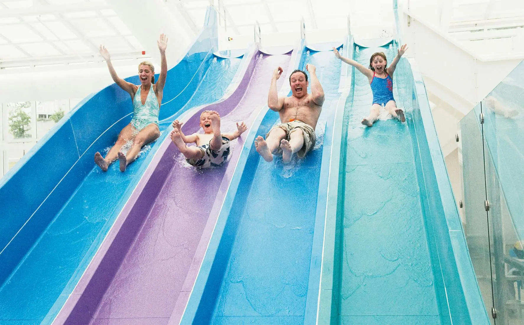 A family of four laughing with arms raised while sliding down indoor multi-lane water slides.