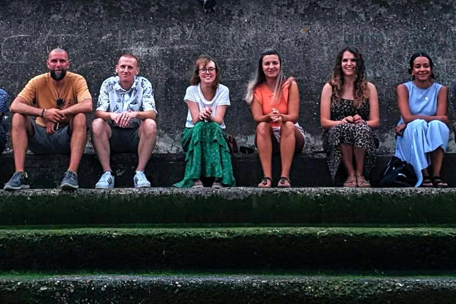 A group of seven people sitting in a row on weathered stone steps against a rough concrete wall.