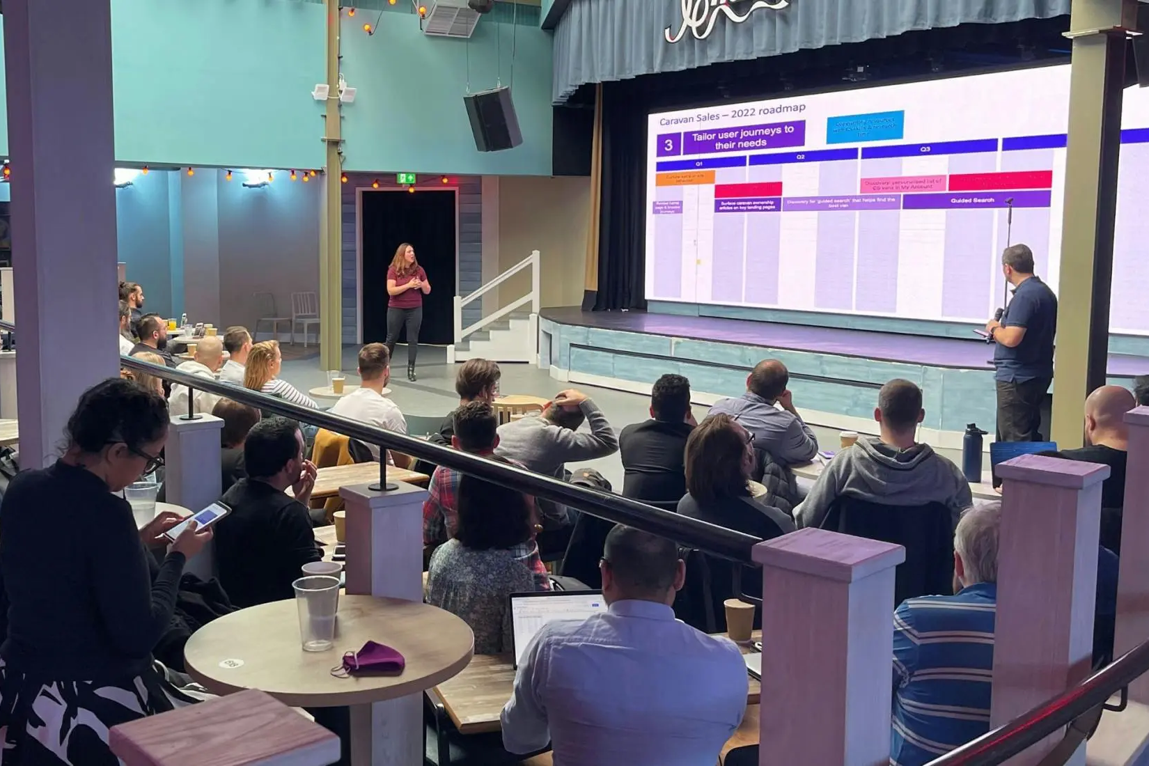 A group of people sitting at tables listening to a speaker presenting a 2022 roadmap on a large screen.