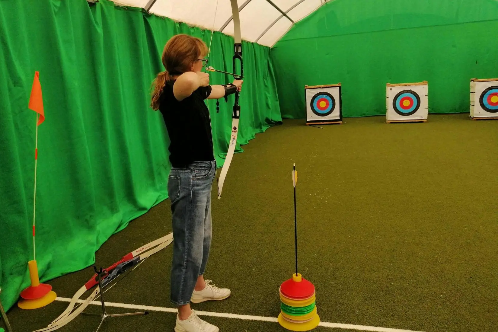 A woman in a black shirt and jeans aiming a recurve bow at target stands in an indoor range.