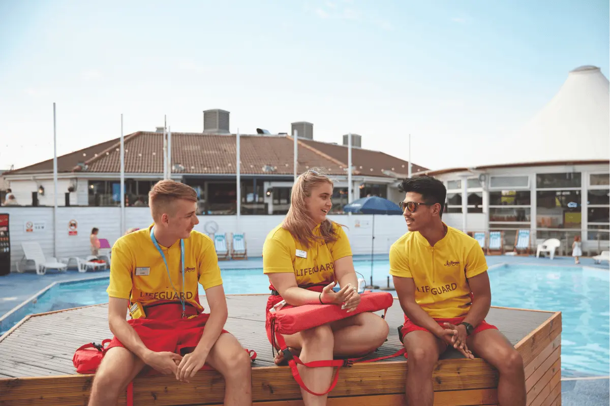 Three lifeguards in yellow shirts and red shorts sitting on a wooden pool deck chatting.