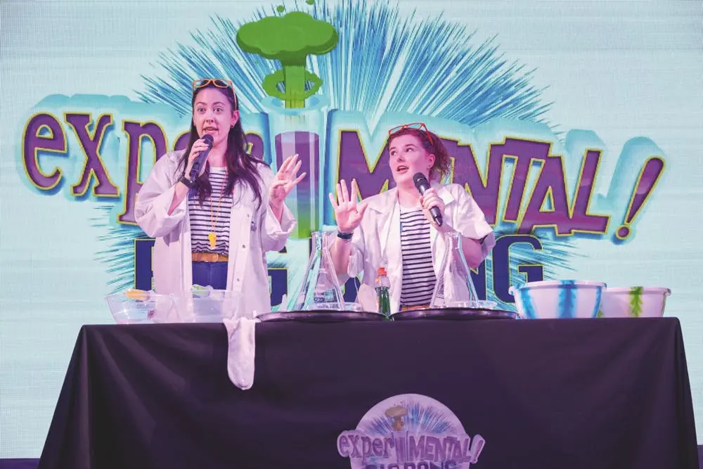 Two presenters in lab coats and striped shirts standing behind a table with science equipment.