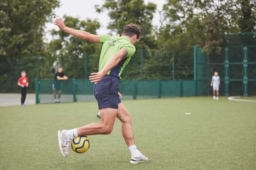 A man in a green activity team shirt kicks a yellow football on an outdoor turf pitch.