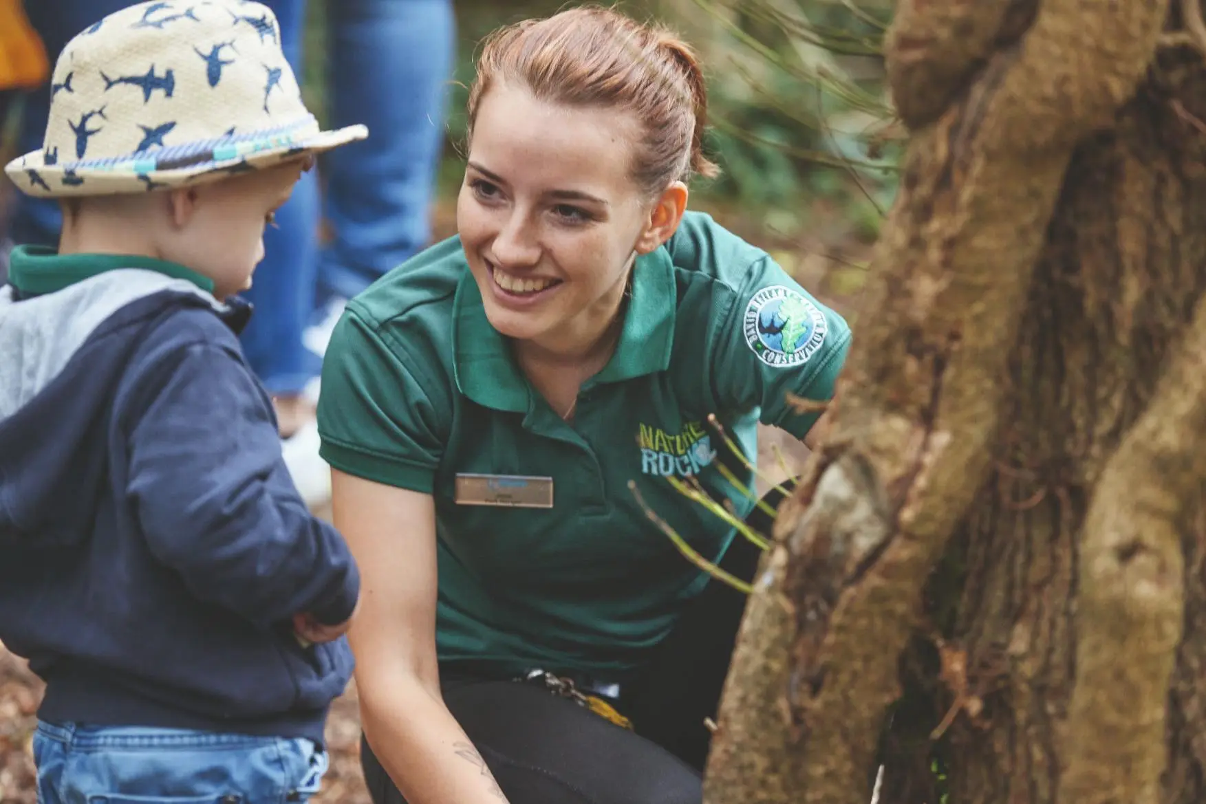 A smiling nature educator interacting with a young boy wearing a shark-patterned hat outdoors.