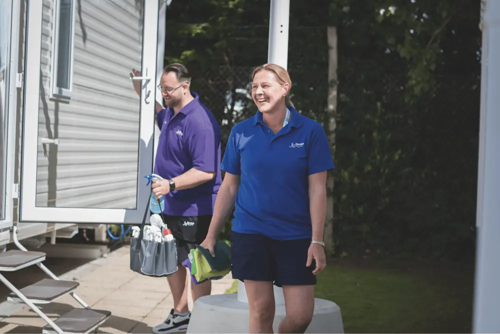A man and a woman in blue uniforms carrying cleaning supplies outside a holiday caravan.