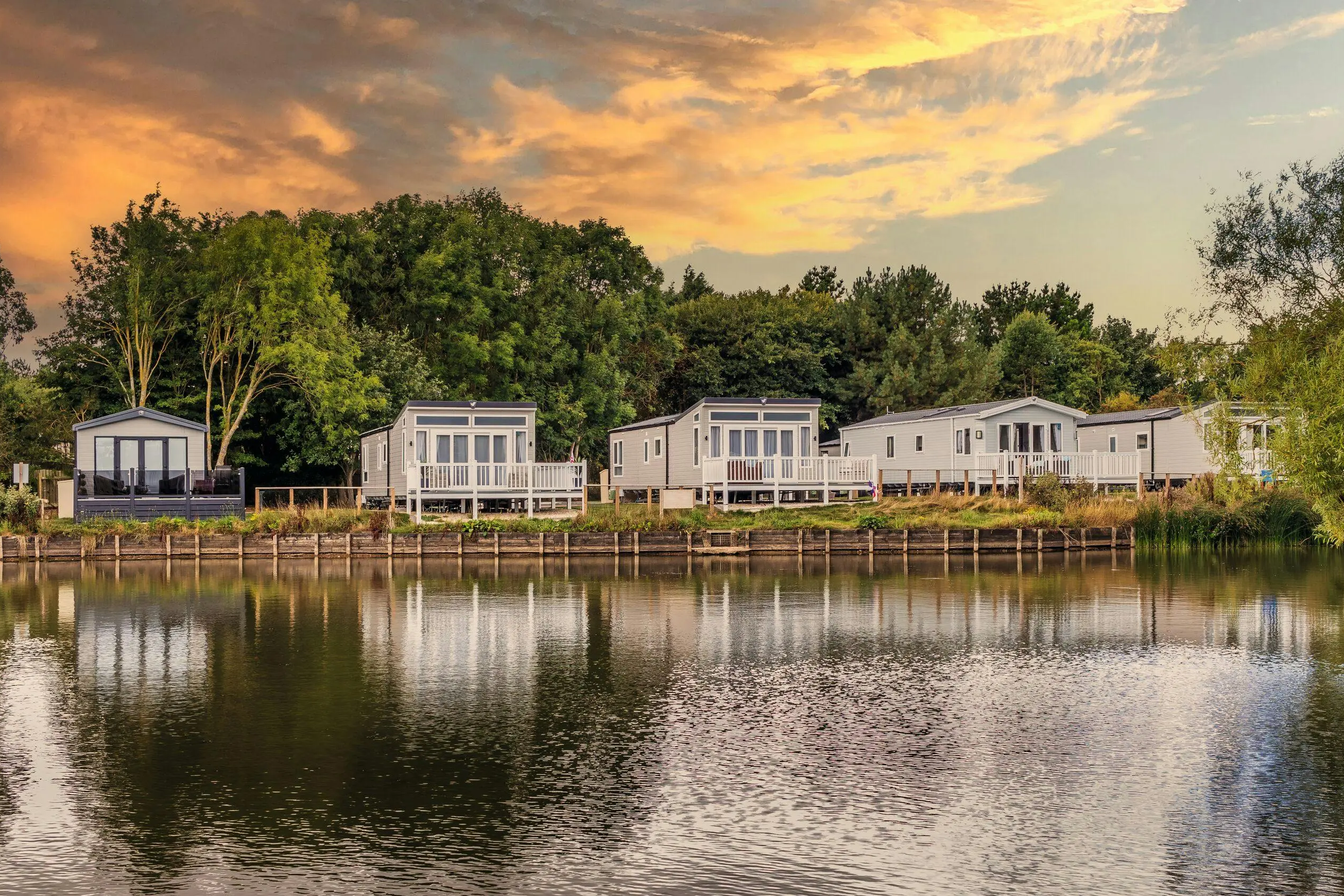 A row of modern static caravans by a tranquil lake at sunset with lush trees in the background.