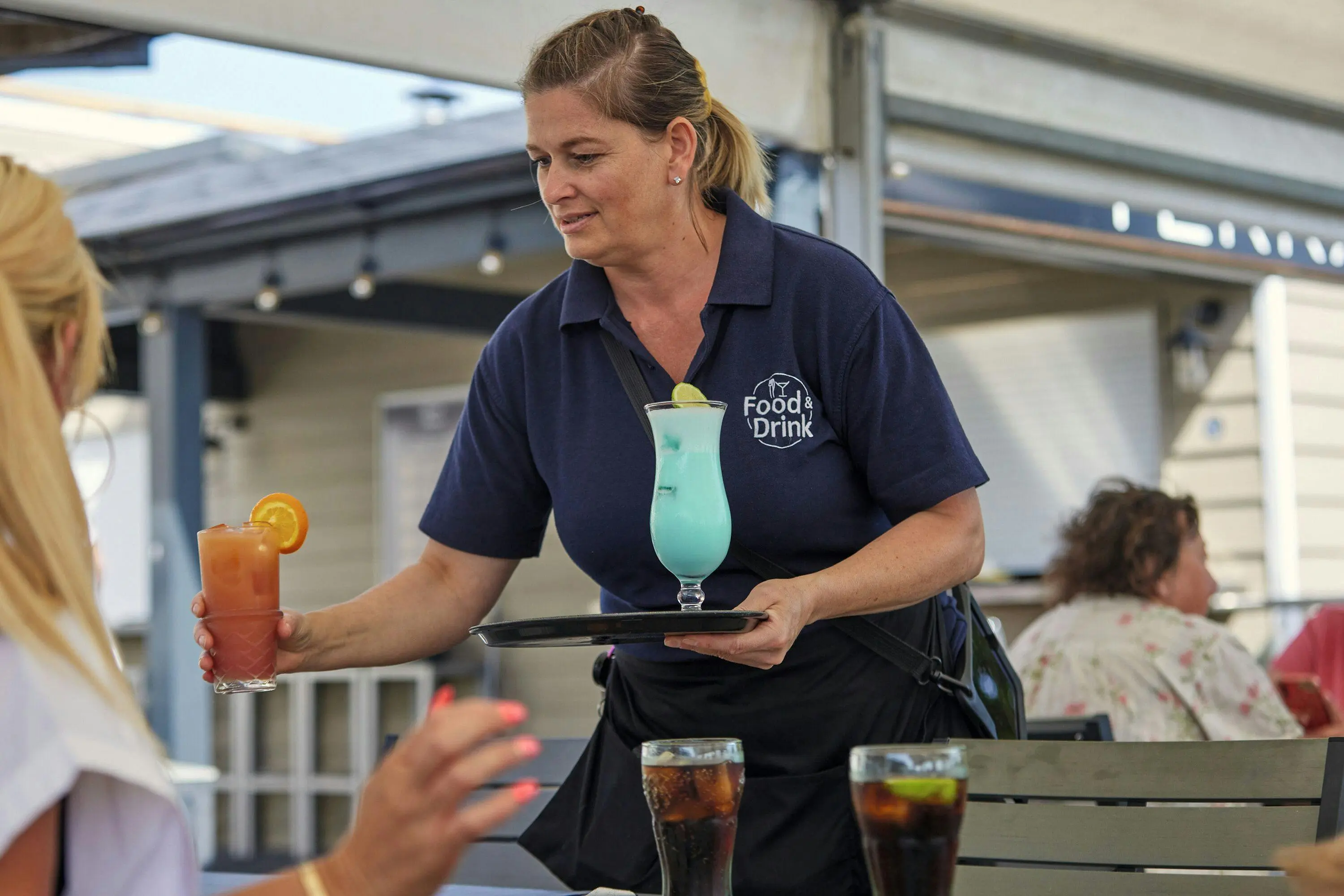 A waitress in a navy uniform serving a blue cocktail and an orange-garnished drink to a customer.