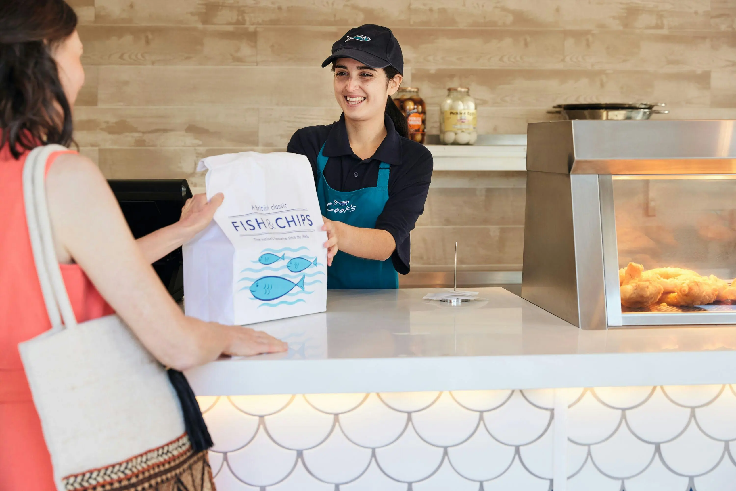 A smiling worker hands a fish and chips takeaway bag to a customer at a shop counter.