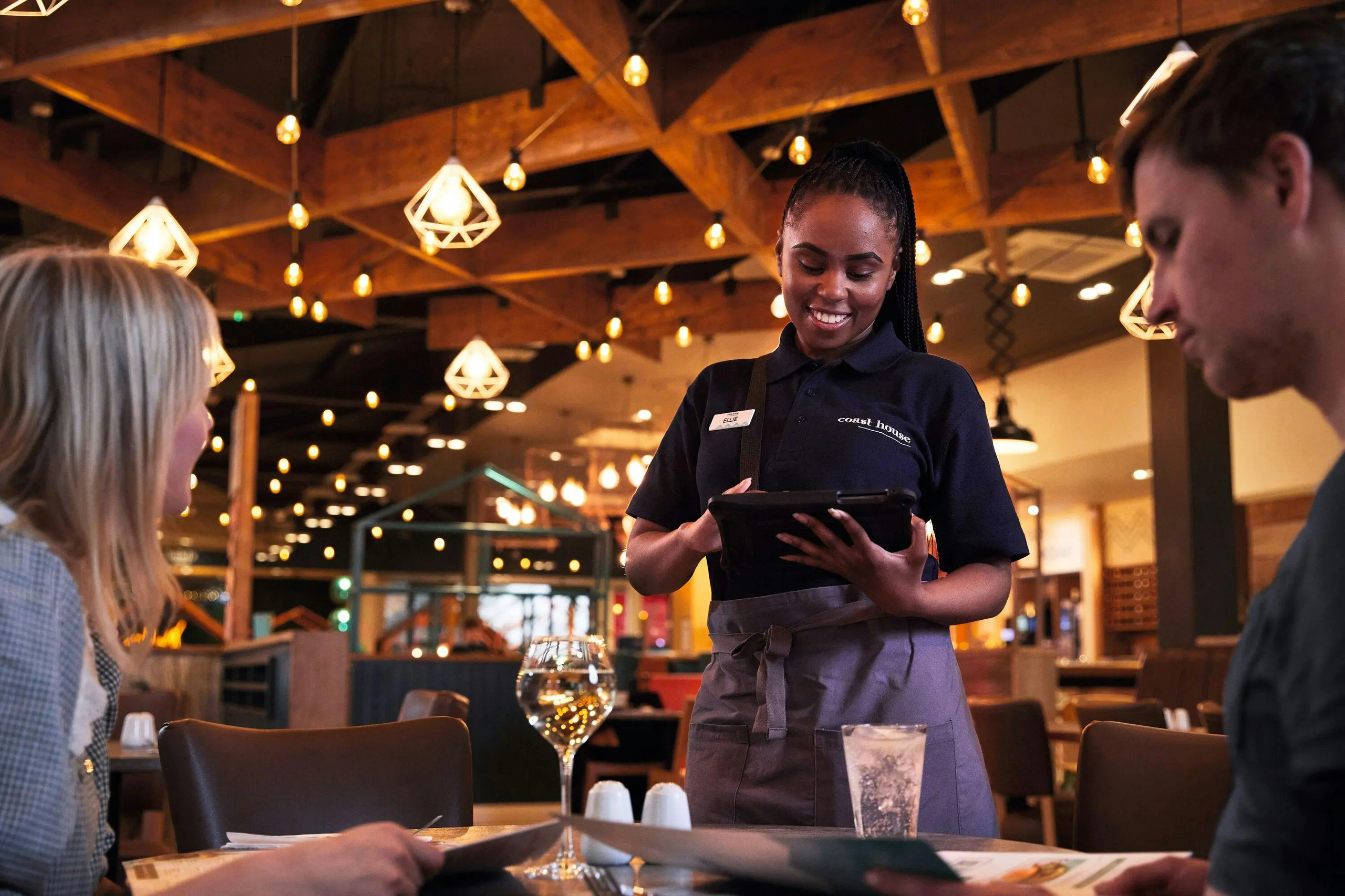 A smiling server at Coast House taking an order on a tablet for customers at a table.