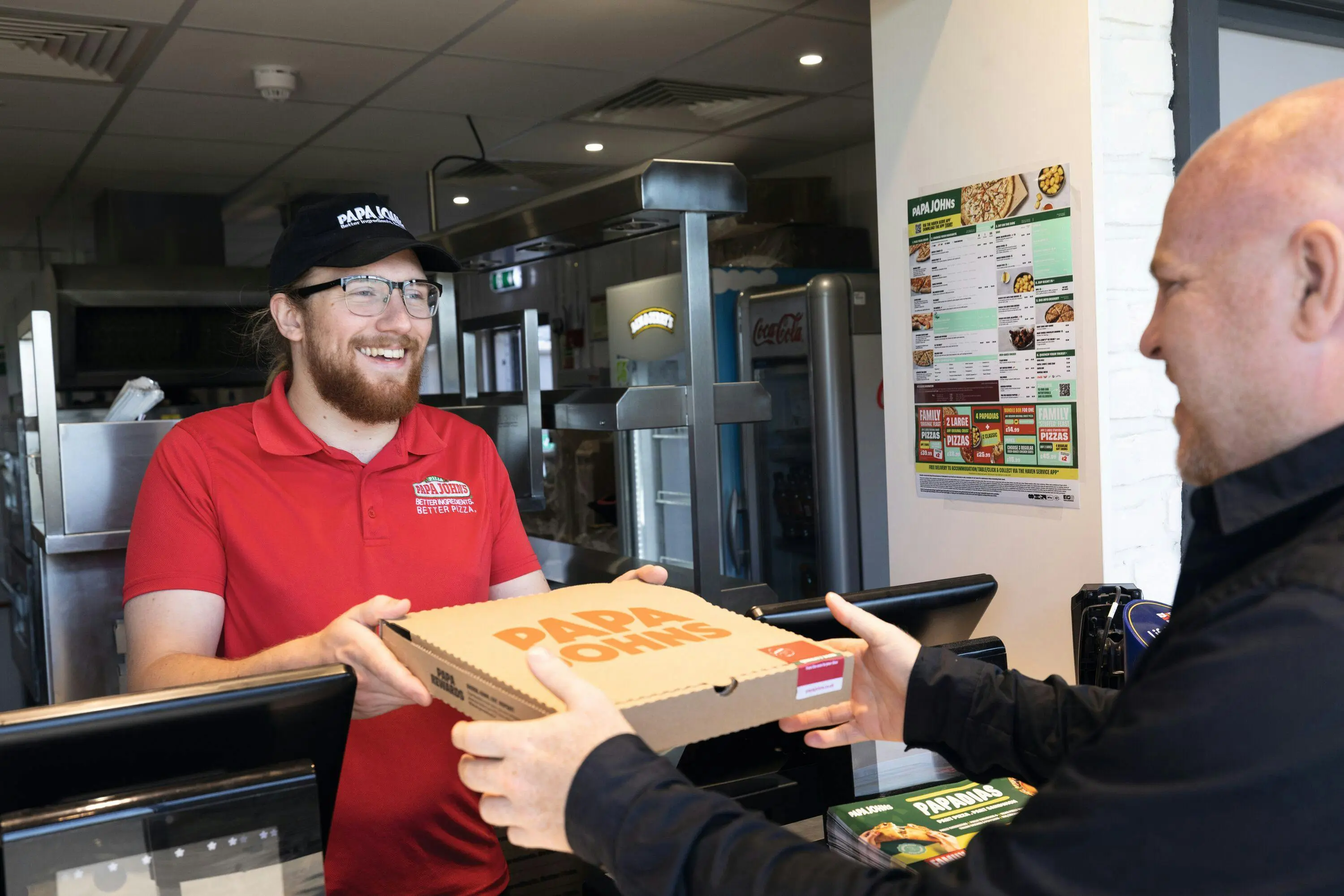 A smiling Papa Johns employee hands a boxed pizza to a customer at the restaurant counter.