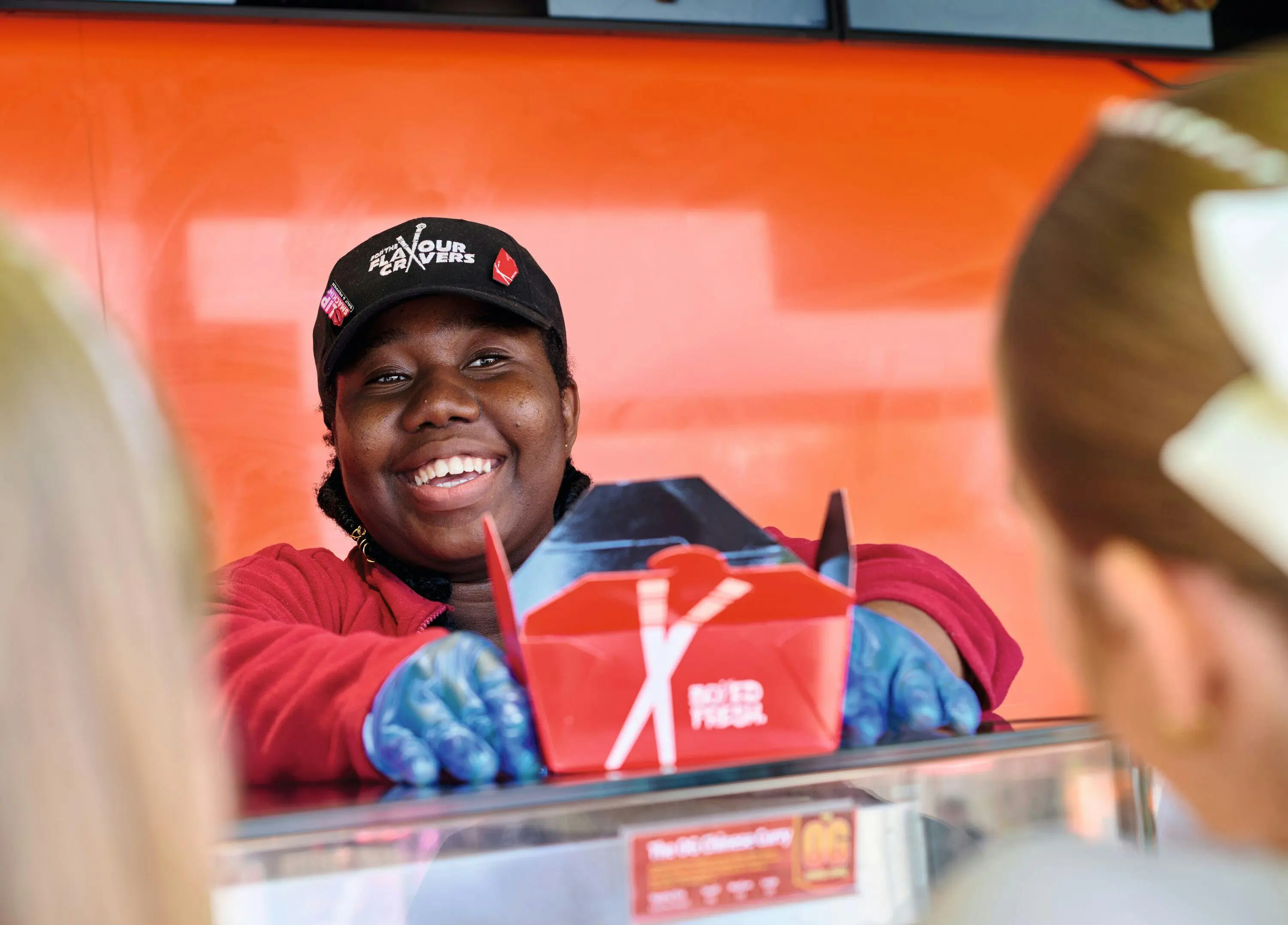 A smiling server in a uniform and gloves handing a red takeout box to customers.