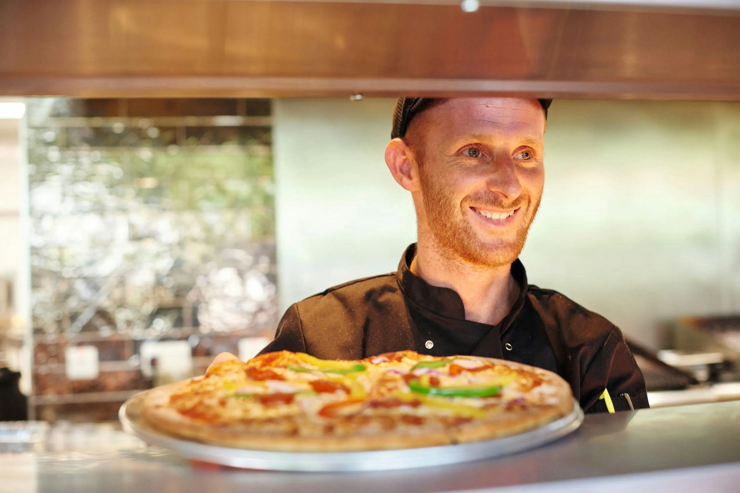 A smiling chef in a black uniform serving a freshly baked pizza in a professional kitchen.