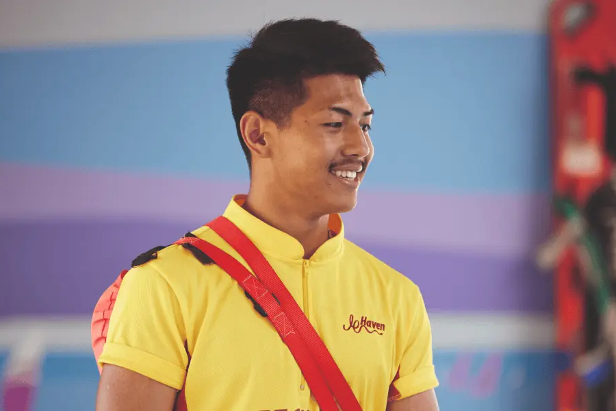 A smiling lifeguard in a yellow uniform with a rescue tube standing in a swimming pool area.