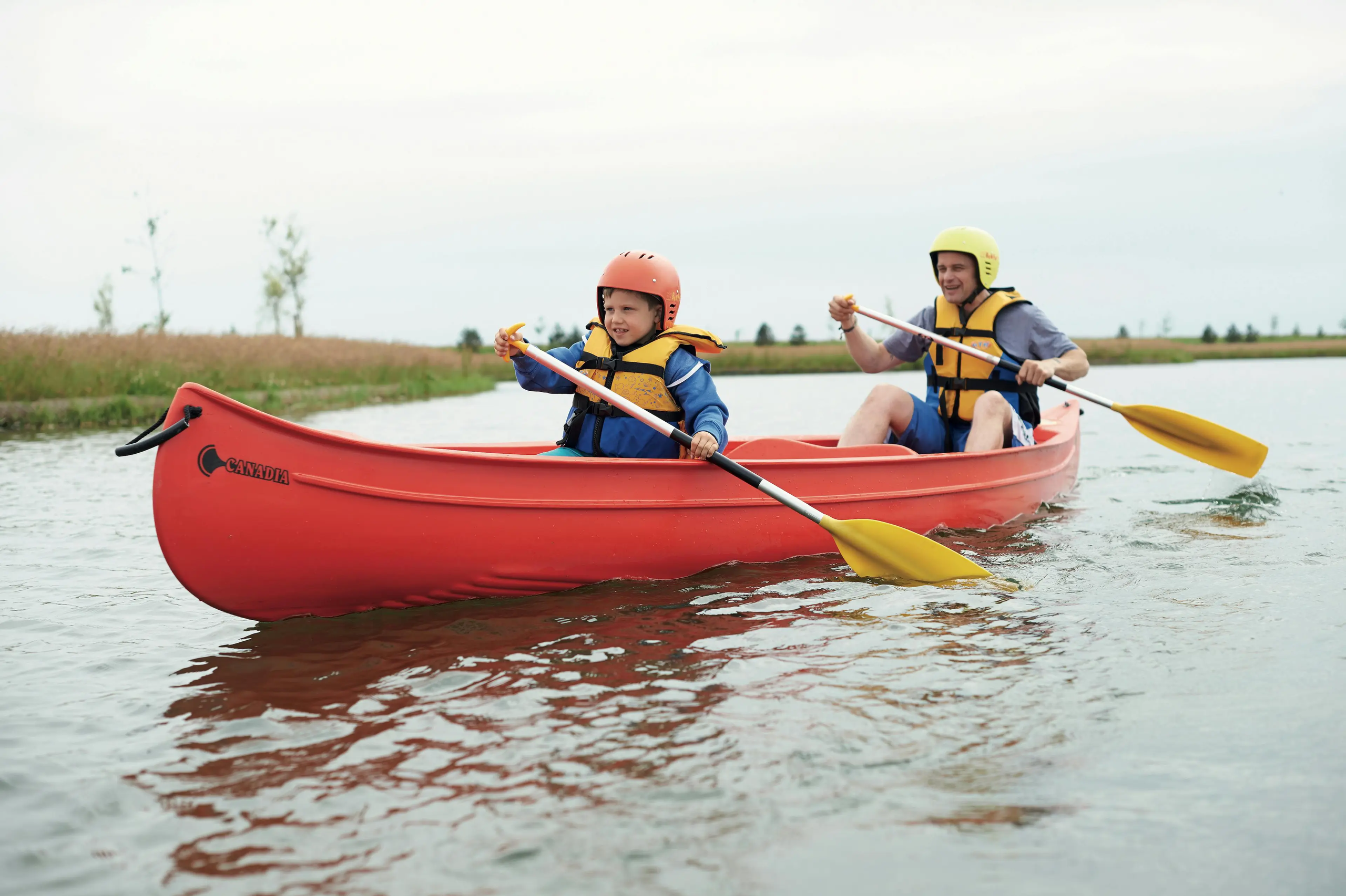 A father and young son in matching life vests and helmets paddling a red canoe on a calm lake.