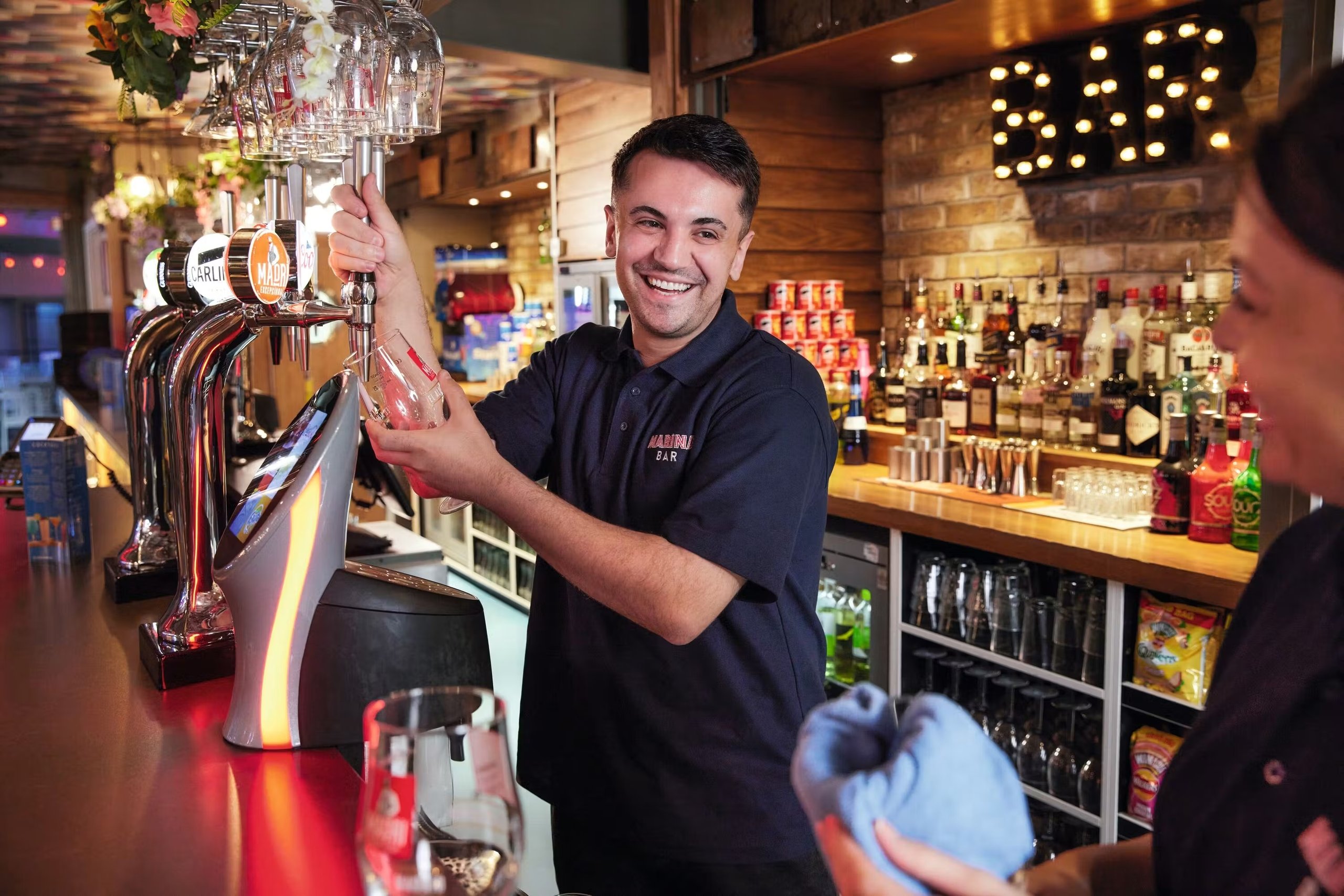 A smiling bartender in a dark polo shirt pours a beer from a tap at a well-stocked bar.
