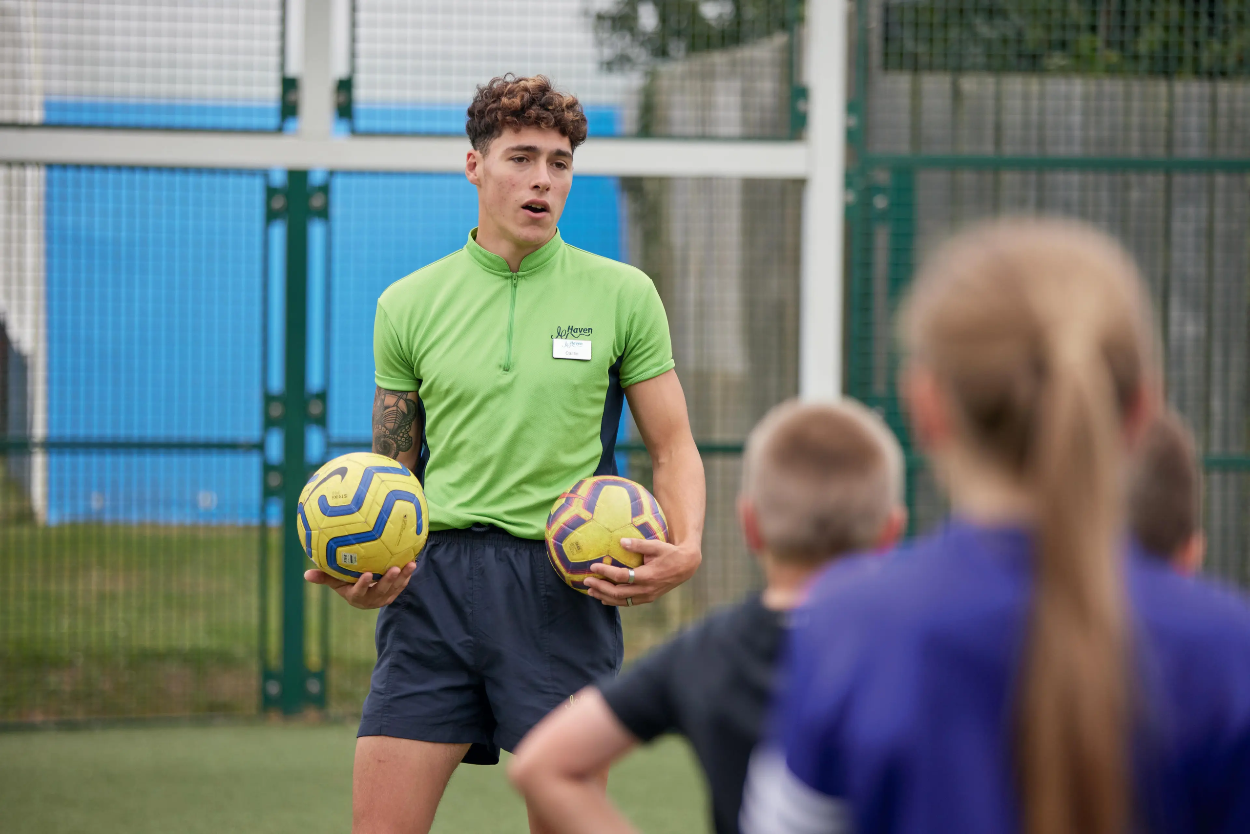 A male soccer coach in a green shirt holding two balls, teaching a group of children on a field.