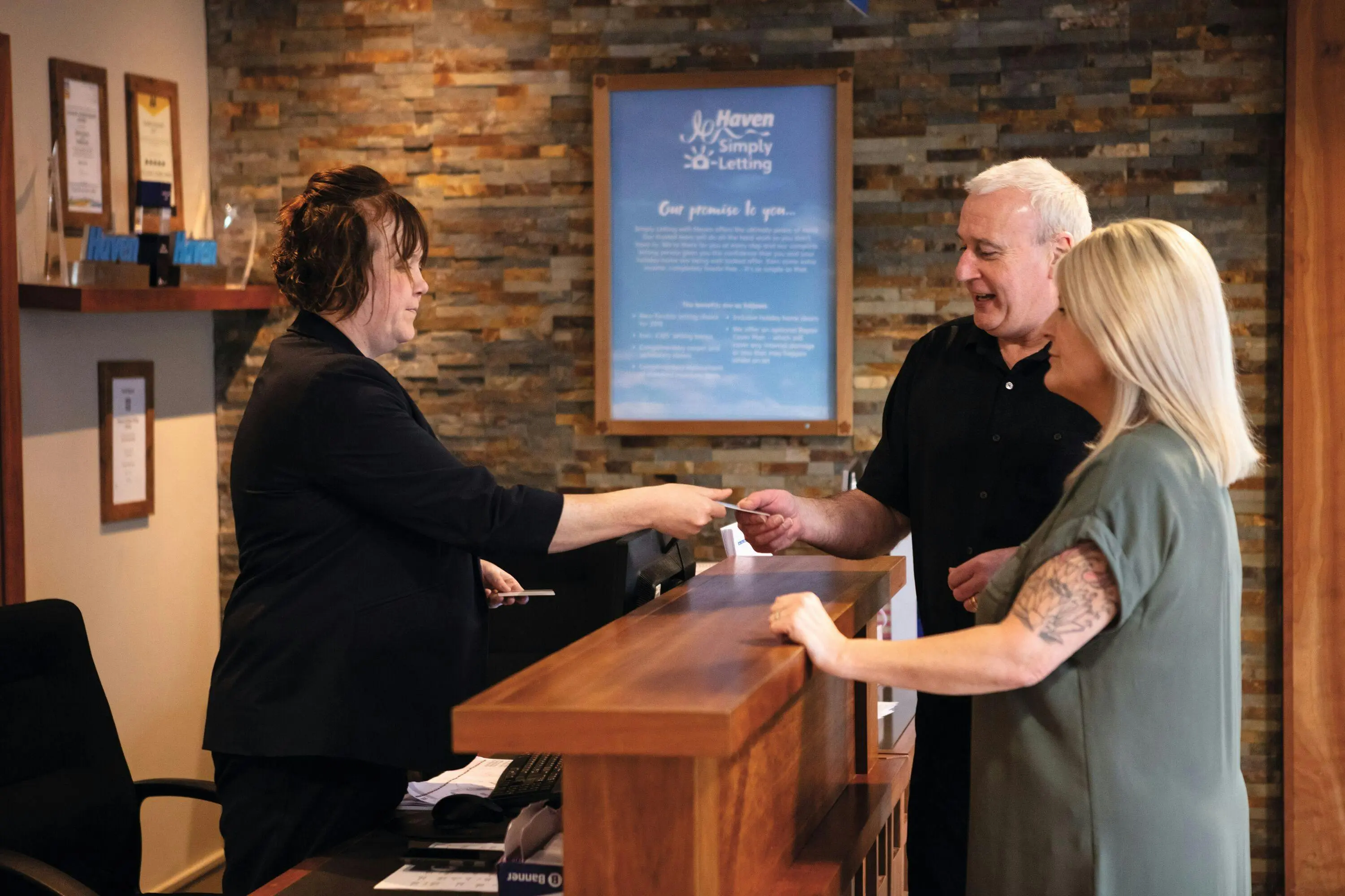 A receptionist hands a card to a couple at a stone-walled office reception desk.