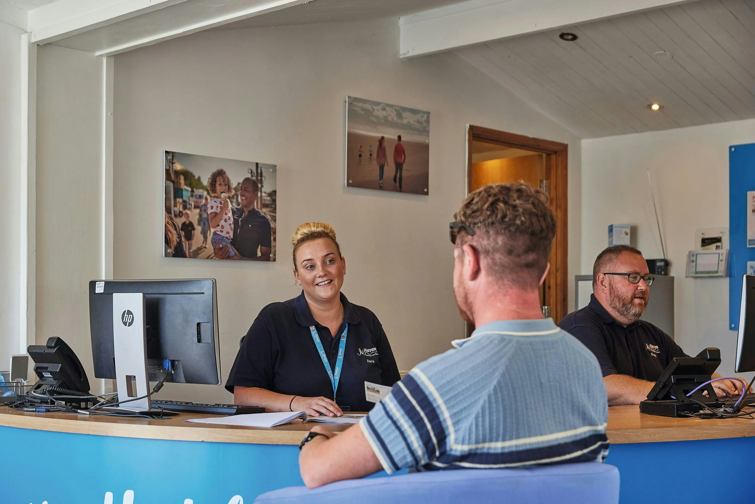 A smiling female employee assisting a male customer at a reception desk while a colleague works.