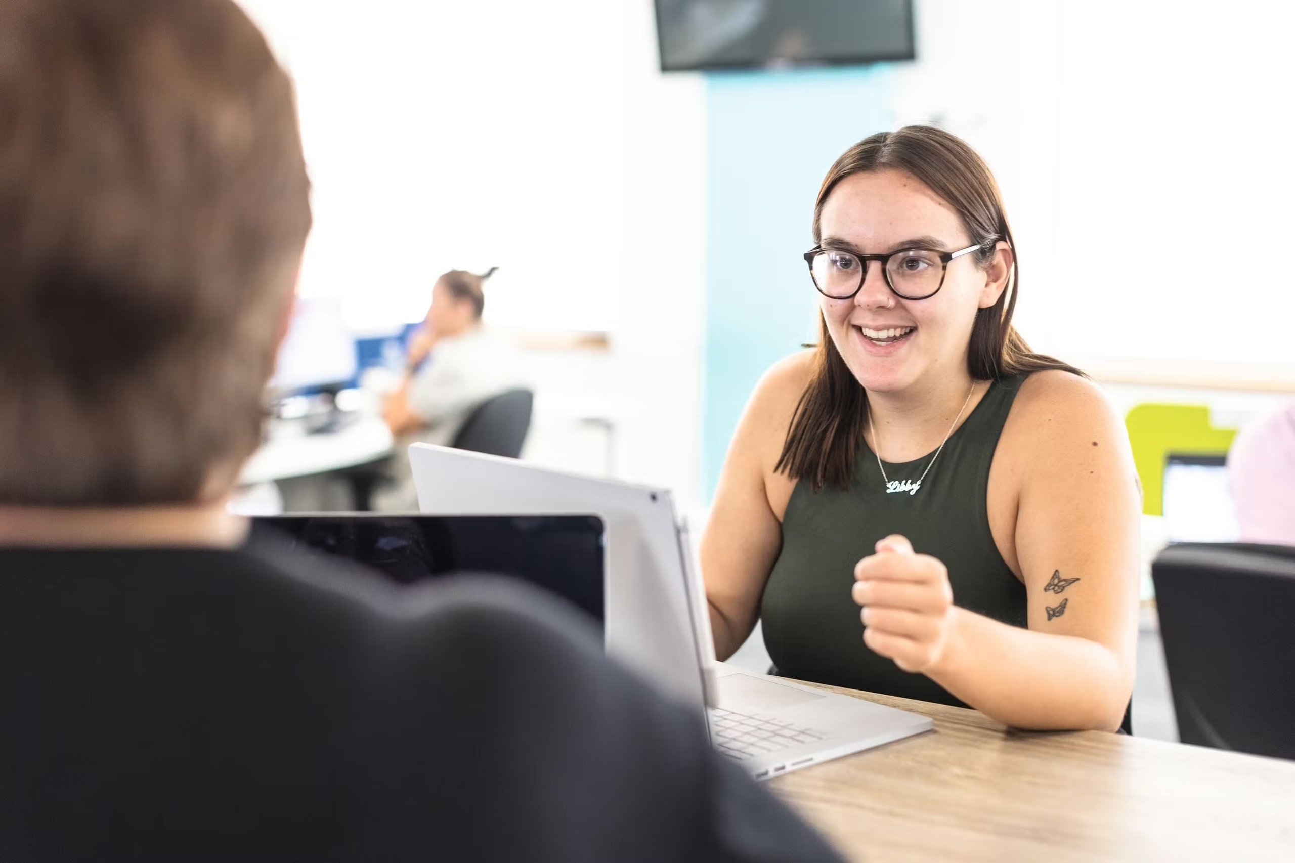 A smiling woman with glasses and a name necklace talking to a colleague in an office setting.