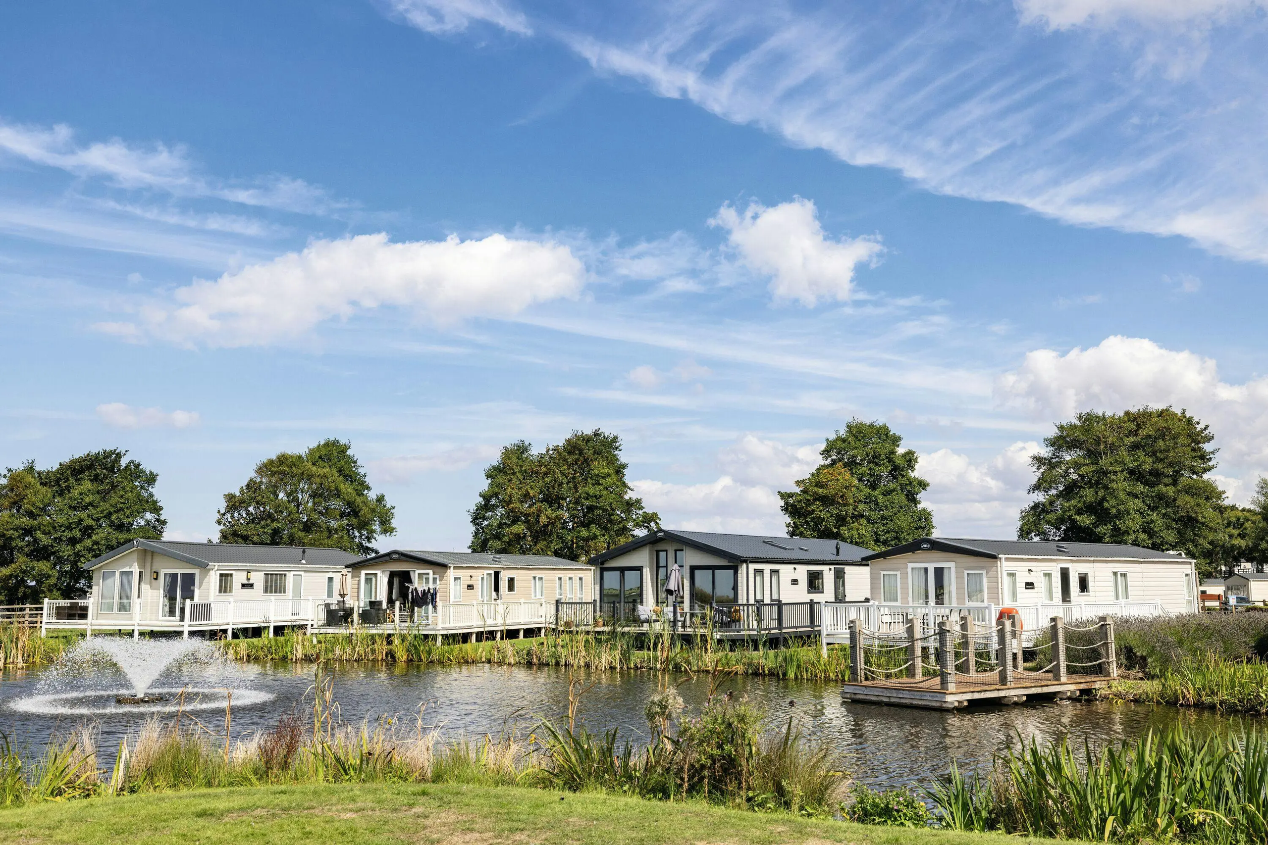 A row of modern static caravans by a pond with a fountain and wooden dock under a blue sky.