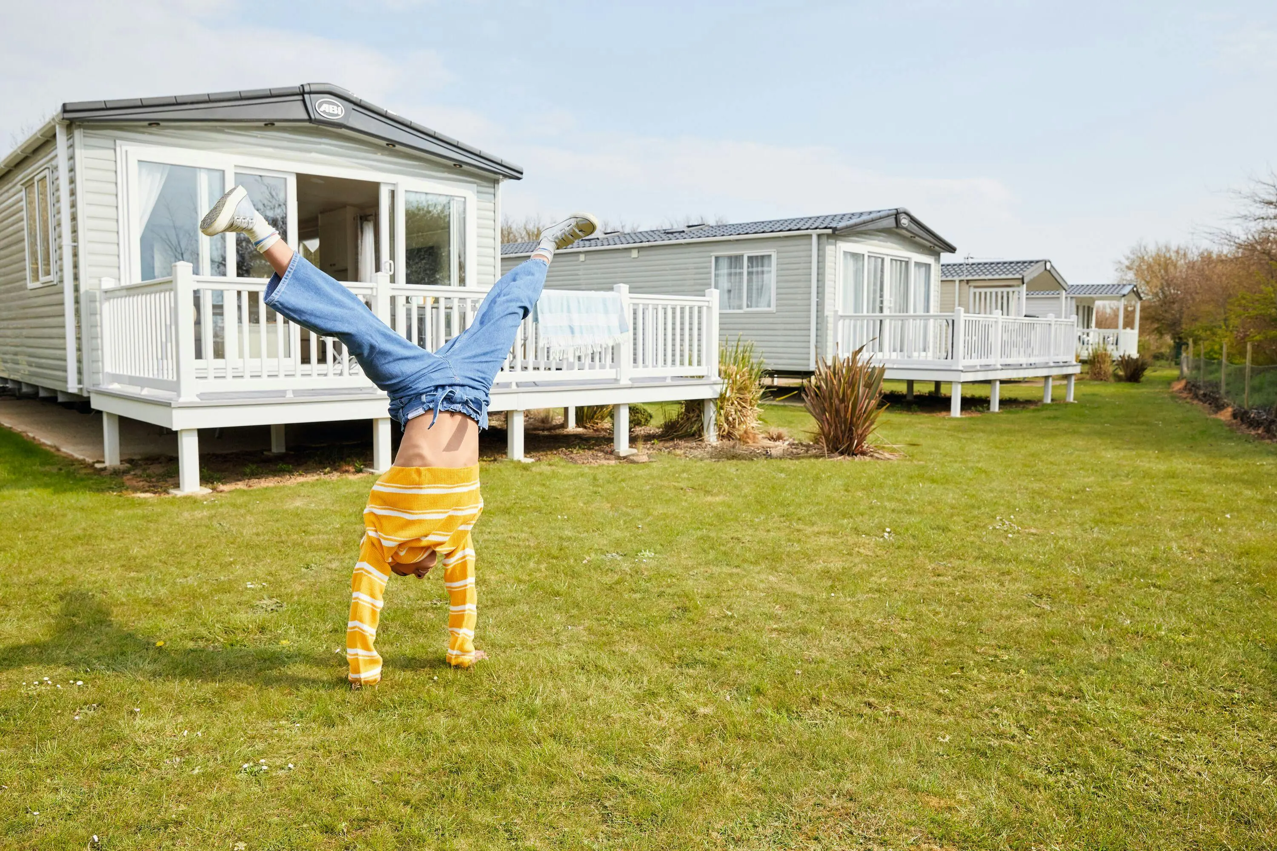 A child in a yellow striped sweater and jeans performs a handstand on the grass near mobile homes.