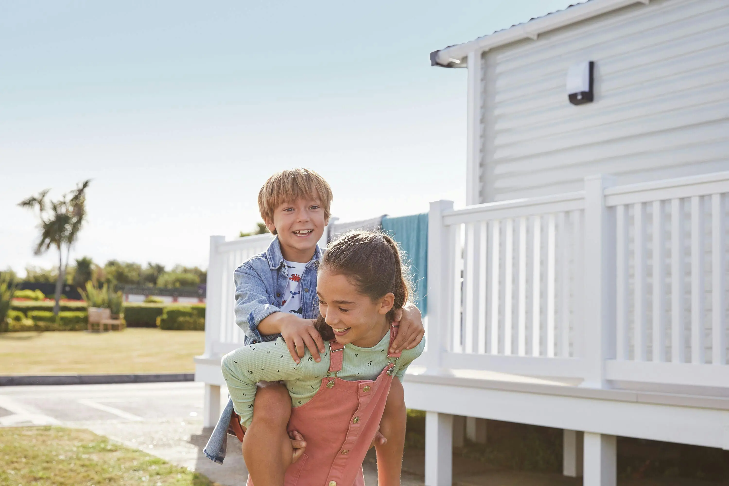 A girl giving a smiling boy a piggyback ride outdoors next to a white holiday home.