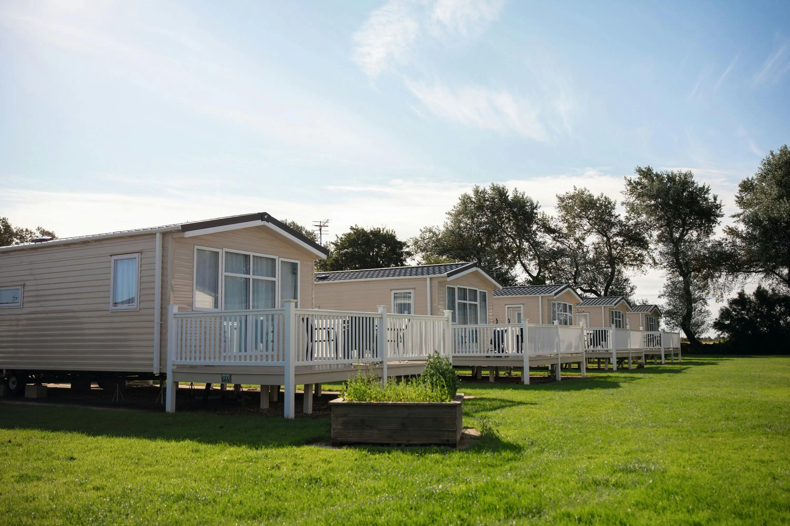 A row of modern static caravans with white wooden decks arranged on a green lawn under a sunny sky.