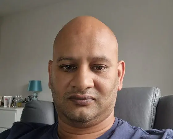 A close-up portrait of a middle-aged bald man with stubble, looking directly at the camera.