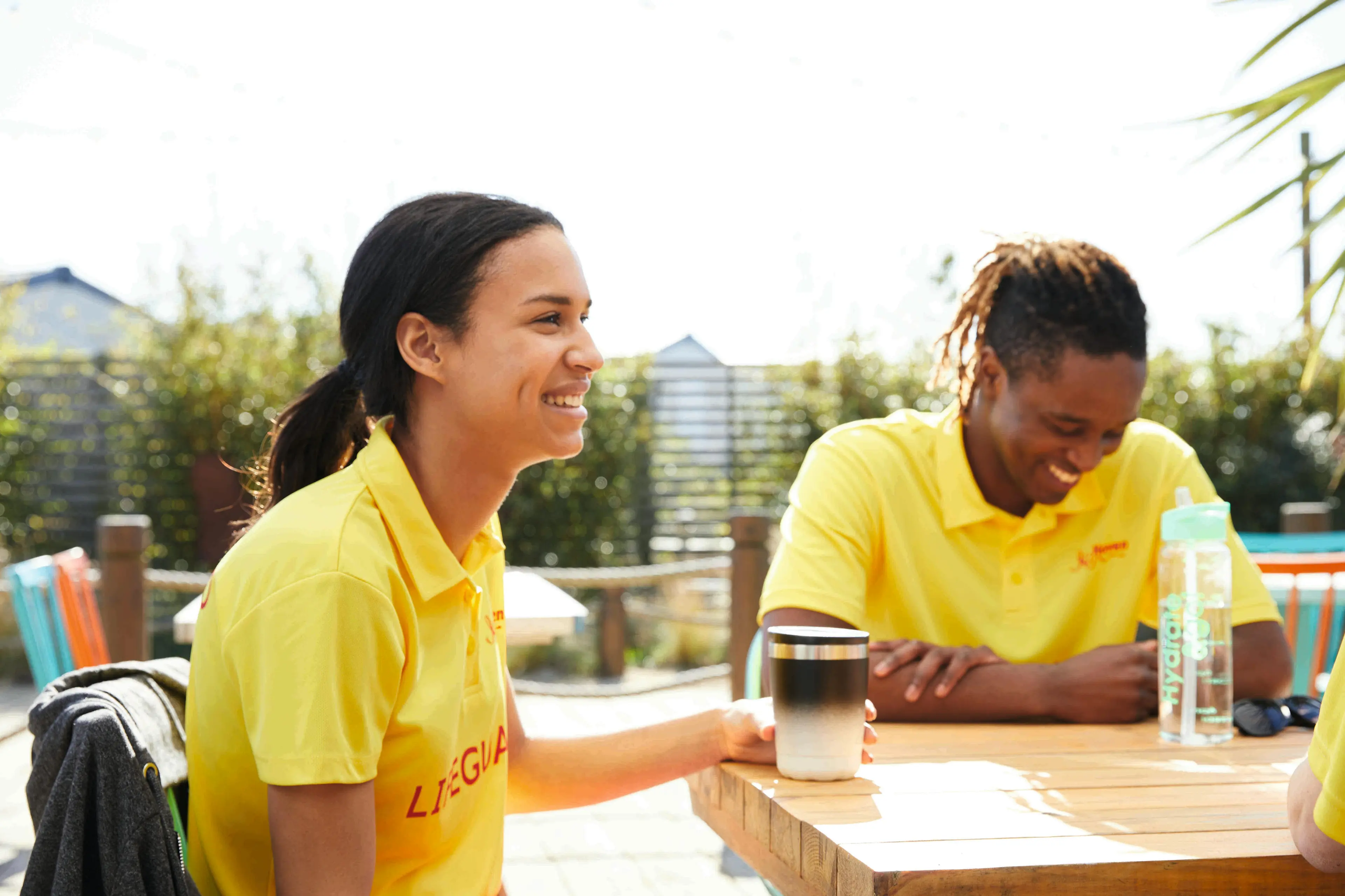 Two smiling lifeguards in yellow uniforms sitting at an outdoor wooden table with drinks.