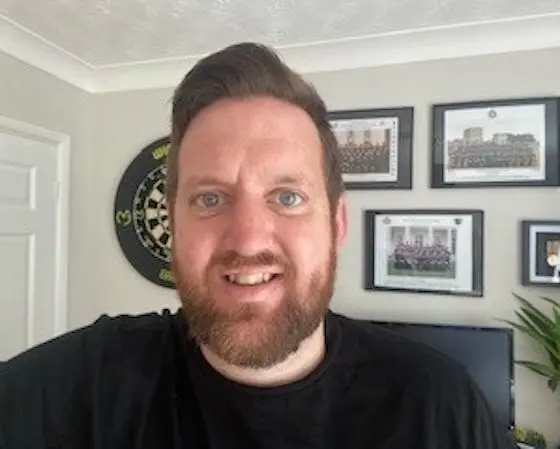 A man with a beard wearing a black shirt, with a dartboard and framed photos on the wall behind him.