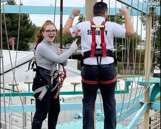 A woman and a security worker wearing safety harnesses on an outdoor high ropes course.