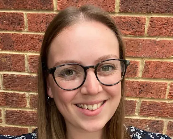 Katherine Lavender smiling in front of a red brick wall, wearing glasses.