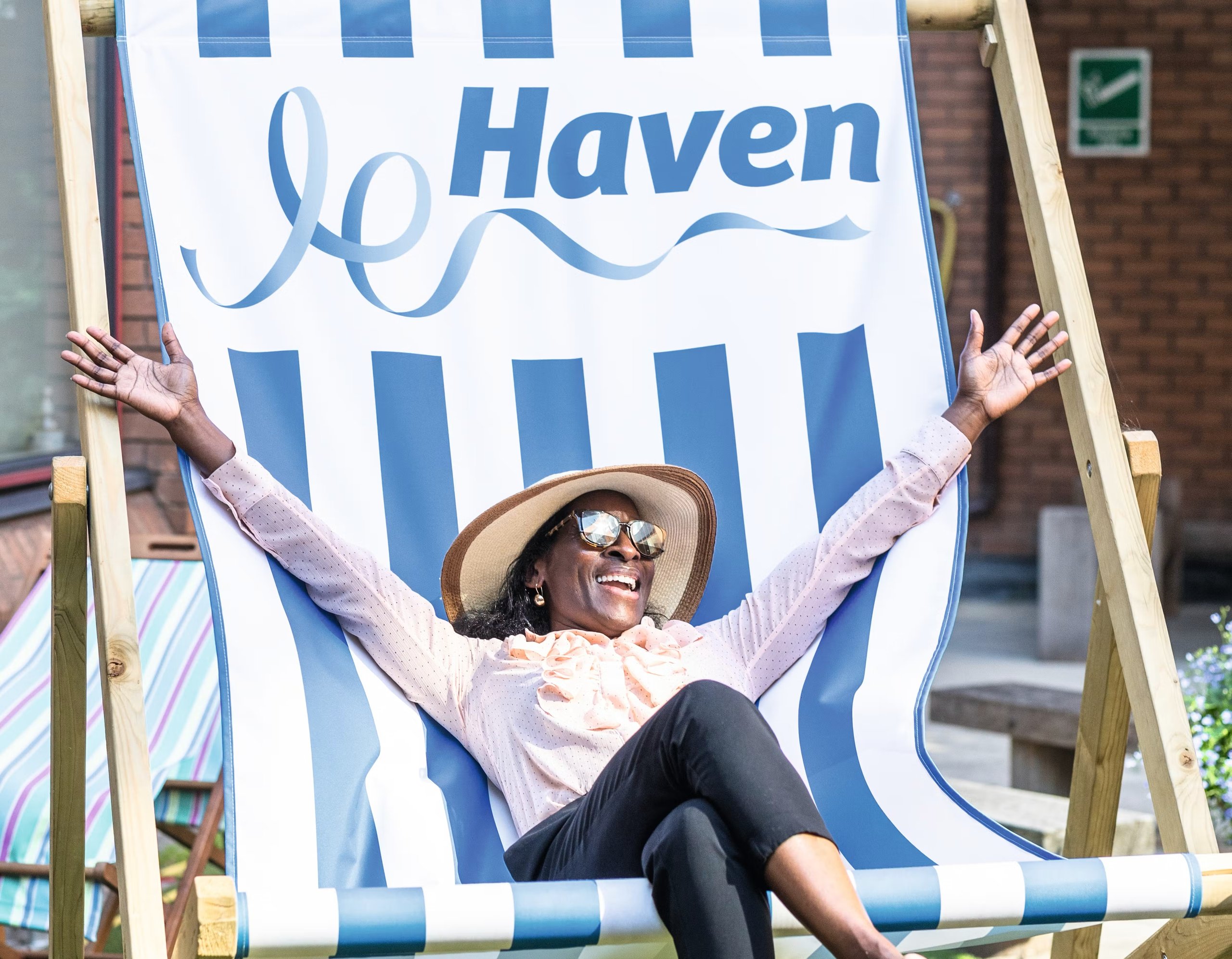 A woman wearing a sun hat and sunglasses sits in a large blue and white striped Haven beach chair.