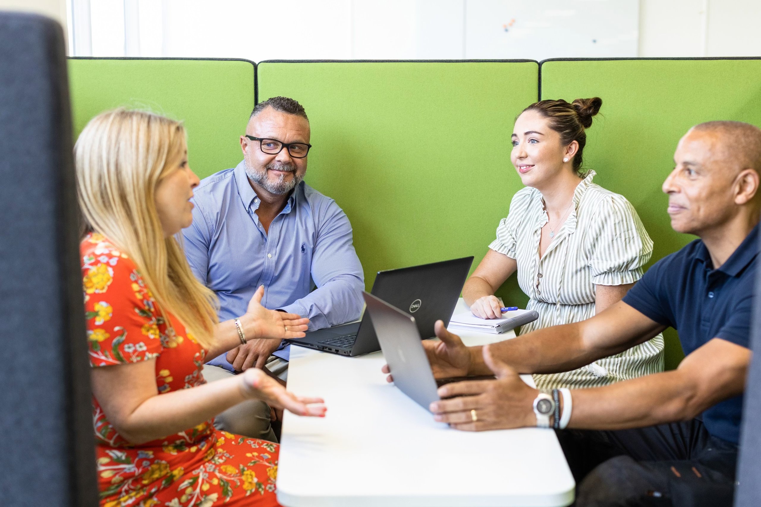 Four diverse professionals sitting at a small table in a green acoustic office pod collaborating.