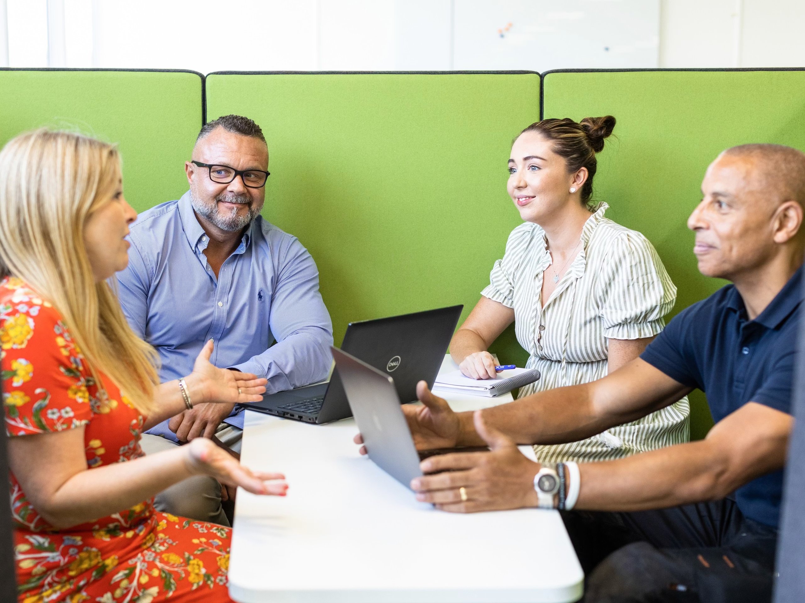 Four diverse professionals sitting at a small table in a green acoustic office pod collaborating.
