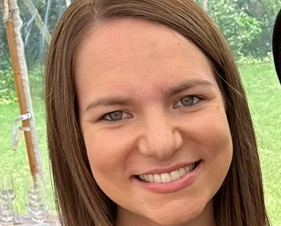 Close-up portrait of a woman with long light brown hair smiling at the camera outdoors.